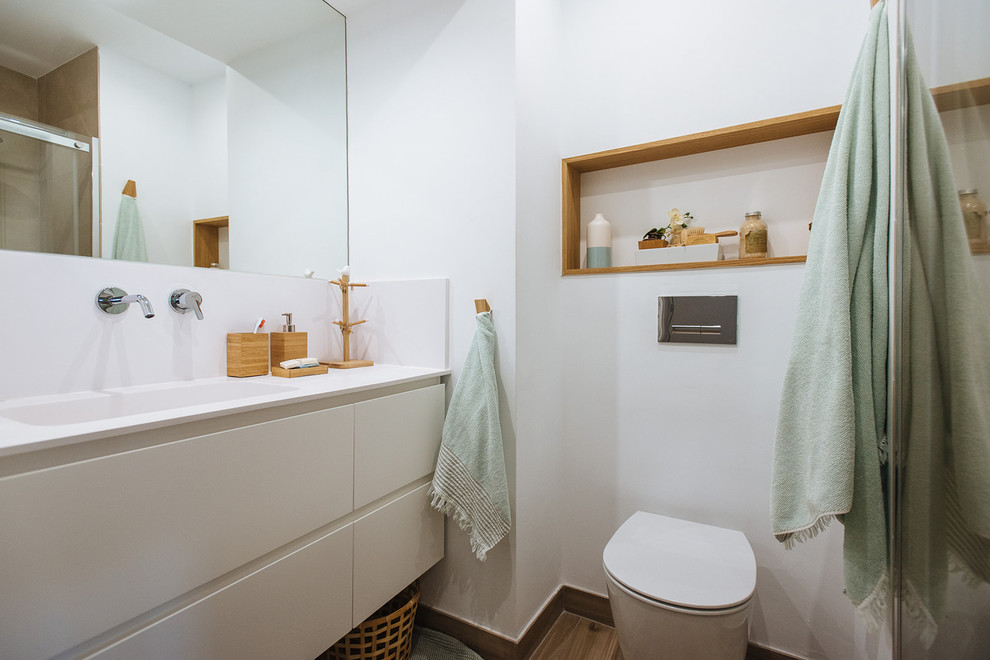 Réalisation d'une salle d'eau design avec un placard à porte plane, des portes de placard blanches, WC suspendus, un mur blanc, un lavabo intégré, un sol marron et un plan de toilette blanc.