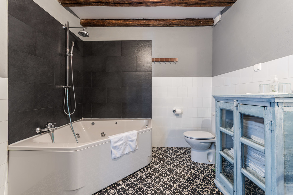 Photo of a medium sized rustic ensuite bathroom in Other with glass-front cabinets, blue cabinets, a hot tub, a shower/bath combination, a two-piece toilet, black and white tiles, ceramic flooring, ceramic tiles, grey walls and a trough sink.