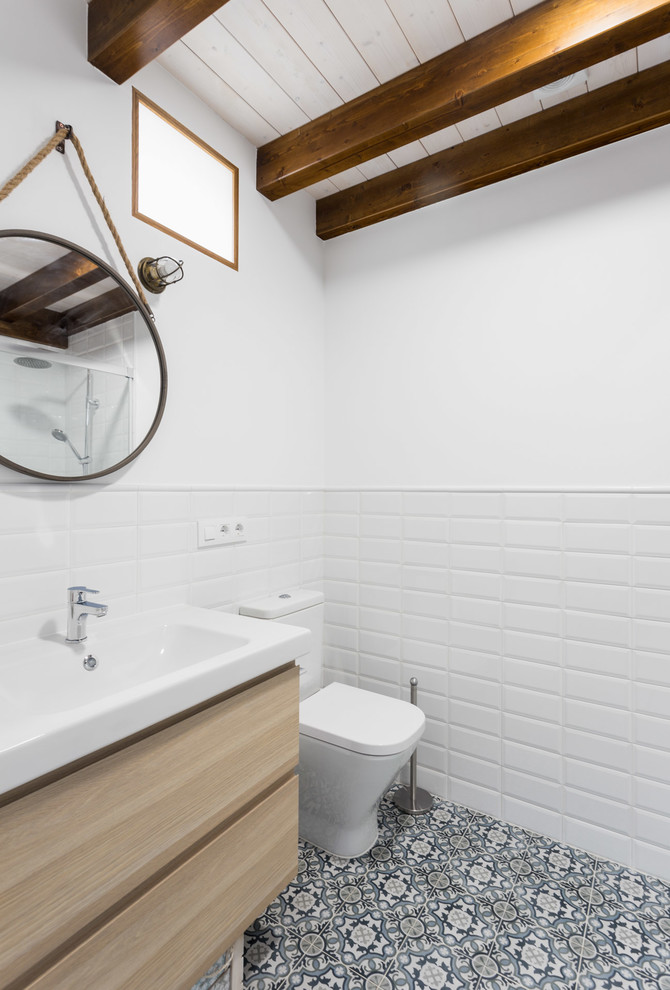 Industriell inredning av ett vit vitt badrum med dusch, med släta luckor, skåp i ljust trä, en toalettstol med hel cisternkåpa, vit kakel, tunnelbanekakel, vita väggar, cementgolv, ett konsol handfat och flerfärgat golv