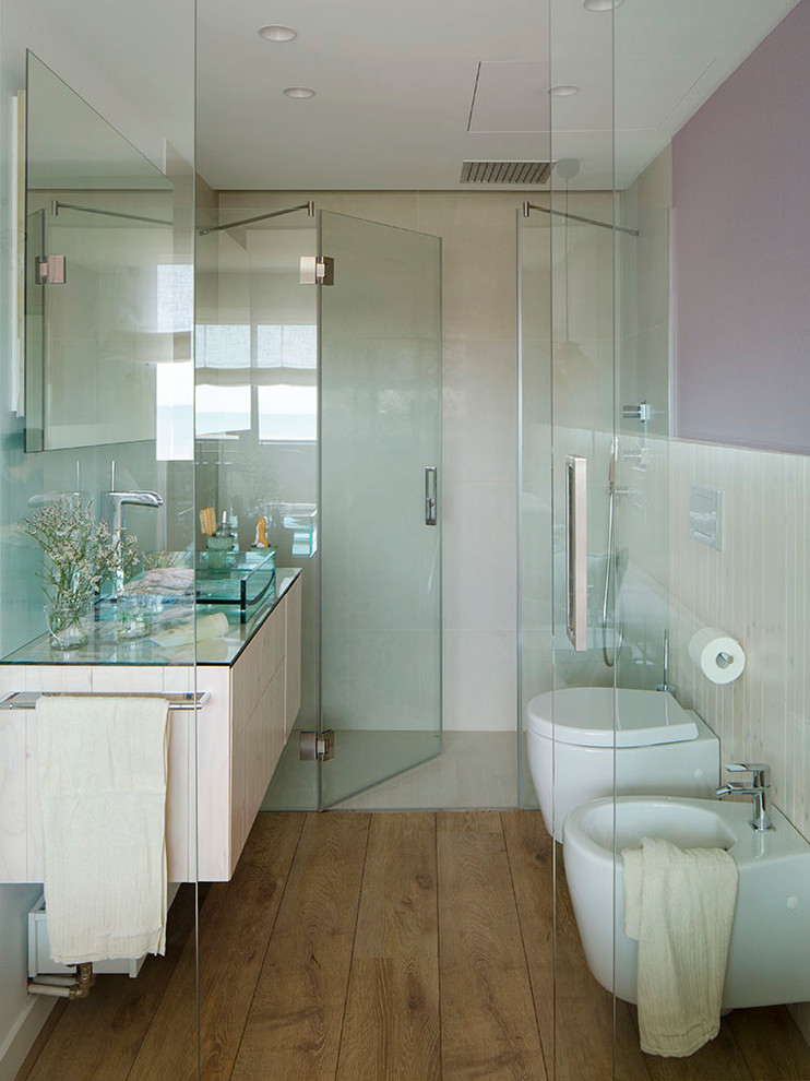 Aménagement d'une salle de bain contemporaine de taille moyenne avec un placard à porte plane, un bidet, des portes de placard beiges, un mur rose, un sol en bois brun, une vasque et un plan de toilette en verre.