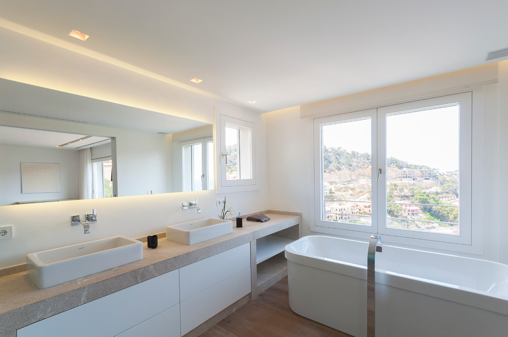 マヨルカ島にある高級な広いコンテンポラリースタイルのおしゃれなマスターバスルーム (フラットパネル扉のキャビネット、白いキャビネット、置き型浴槽、白い壁、無垢フローリング、ベッセル式洗面器、ライムストーンの洗面台) の写真
