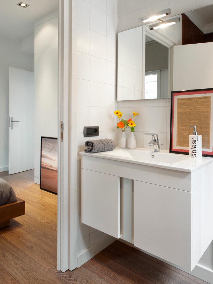 Idée de décoration pour une salle d'eau design de taille moyenne avec un placard à porte plane, des portes de placard blanches, un carrelage blanc, un mur blanc, un sol en bois brun et un lavabo suspendu.