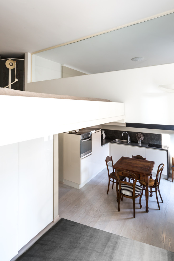 Kleine Moderne Wohnküche in L-Form mit flächenbündigen Schrankfronten, weißen Schränken, Küchenrückwand in Schwarz, hellem Holzboden und grauem Boden in Sonstige