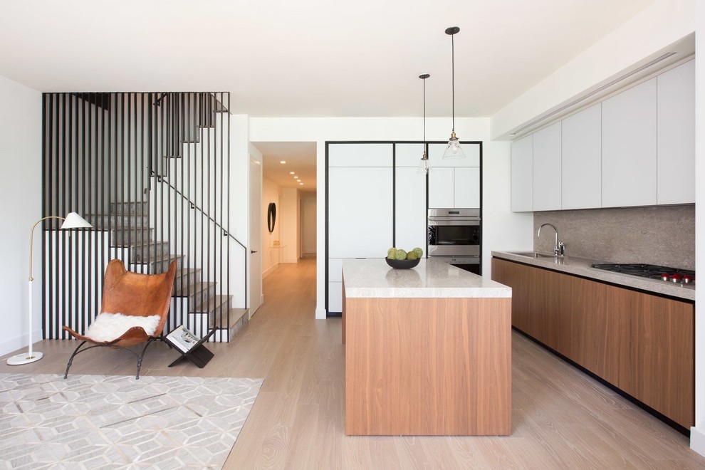 This is an example of a modern open plan kitchen in New York with a submerged sink, flat-panel cabinets, white cabinets, grey splashback, stainless steel appliances, light hardwood flooring, an island and grey worktops.