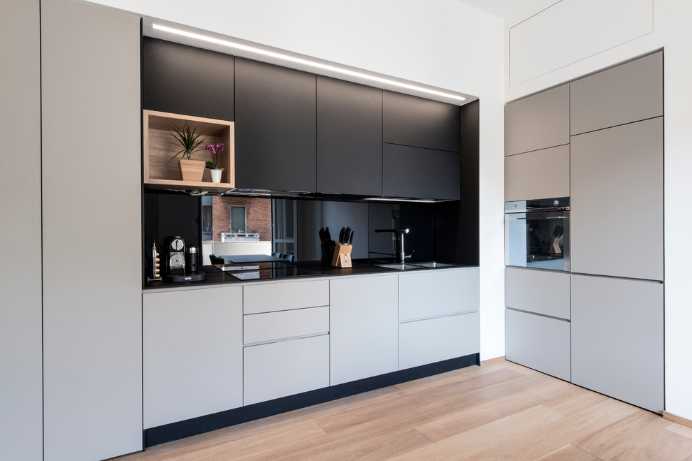 Inspiration for a medium sized contemporary l-shaped kitchen in Milan with a double-bowl sink, flat-panel cabinets, laminate countertops, black splashback, glass sheet splashback, stainless steel appliances, light hardwood flooring, no island and grey cabinets.