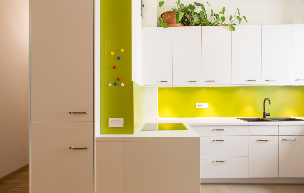 Bild på ett funkis vit vitt l-kök, med en dubbel diskho, släta luckor, vita skåp, grönt stänkskydd och beiget golv