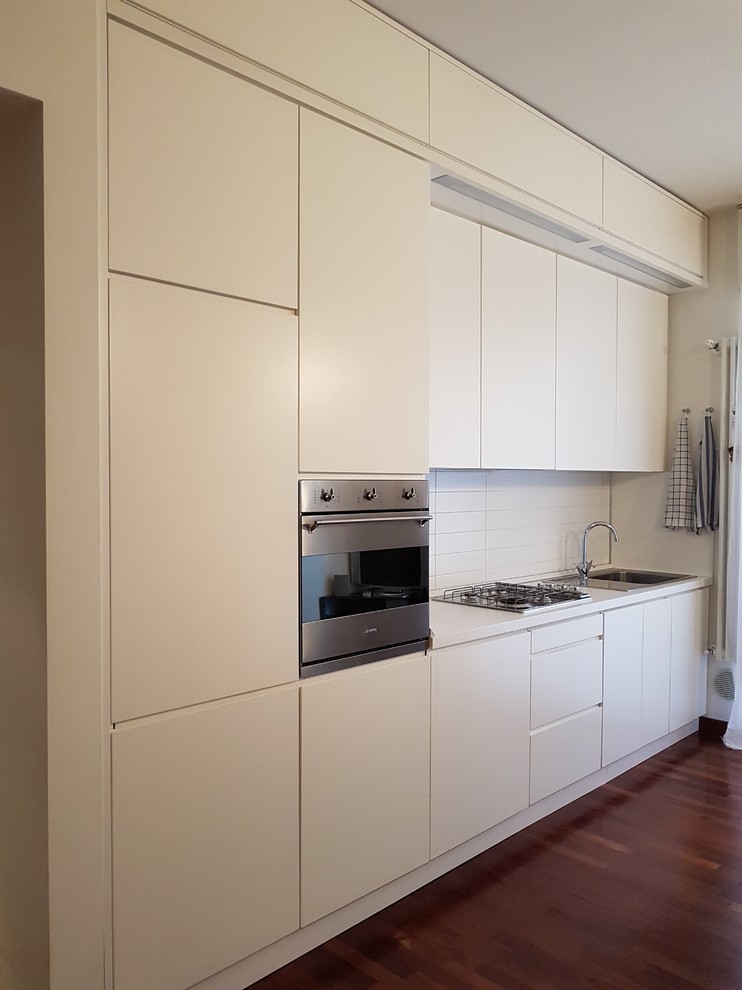 Small contemporary single-wall open plan kitchen in Other with a built-in sink, flat-panel cabinets, white cabinets, laminate countertops, white splashback, ceramic splashback, stainless steel appliances, medium hardwood flooring and no island.