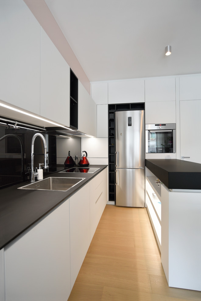 Small contemporary u-shaped open plan kitchen in Milan with a built-in sink, black splashback, light hardwood flooring, a breakfast bar, black worktops, flat-panel cabinets and white cabinets.
