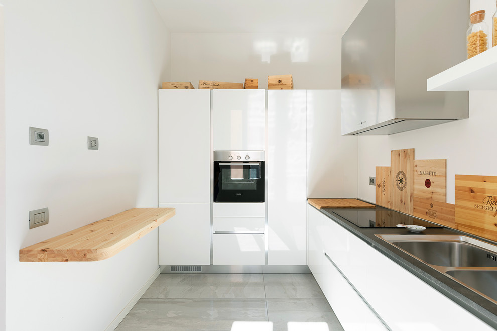 На фото: отдельная, угловая кухня в белых тонах с отделкой деревом в современном стиле с двойной мойкой, плоскими фасадами, белыми фасадами, черной техникой, полуостровом, серым полом и серой столешницей с