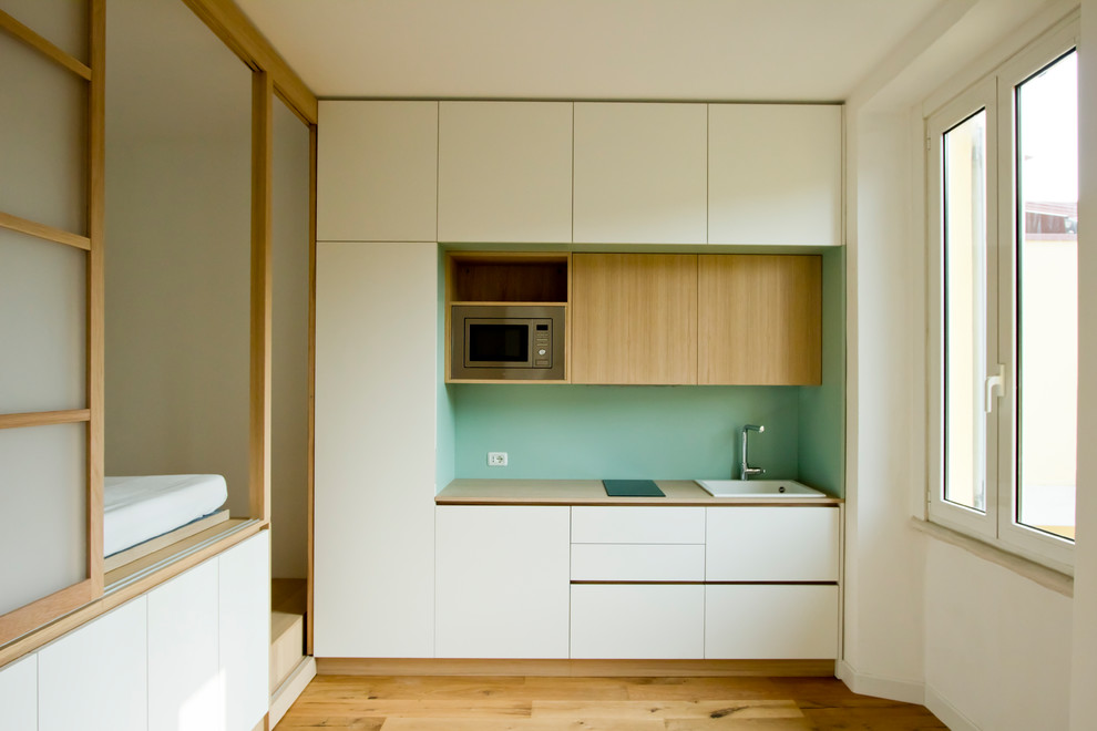 Minimalistisk inredning av ett litet brun linjärt brunt kök med öppen planlösning, med en nedsänkt diskho, släta luckor, vita skåp, träbänkskiva, grönt stänkskydd, stänkskydd i trä, rostfria vitvaror, ljust trägolv och brunt golv