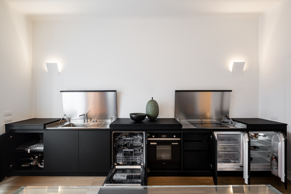 Einzeilige Moderne Wohnküche ohne Insel mit Waschbecken, schwarzen Schränken, Edelstahl-Arbeitsplatte, Küchengeräten aus Edelstahl, hellem Holzboden, braunem Boden und schwarzer Arbeitsplatte in Mailand