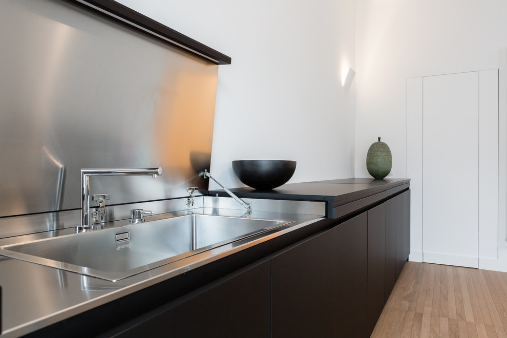 Bild på ett funkis svart linjärt svart kök och matrum, med en enkel diskho, svarta skåp, bänkskiva i rostfritt stål, rostfria vitvaror, ljust trägolv och brunt golv