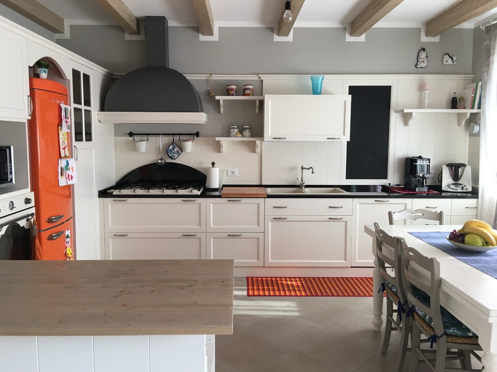 Lantlig inredning av ett kök, med en rustik diskho, vita skåp, granitbänkskiva, svart stänkskydd, stänkskydd i skiffer, vita vitvaror, klinkergolv i porslin, en halv köksö och beiget golv