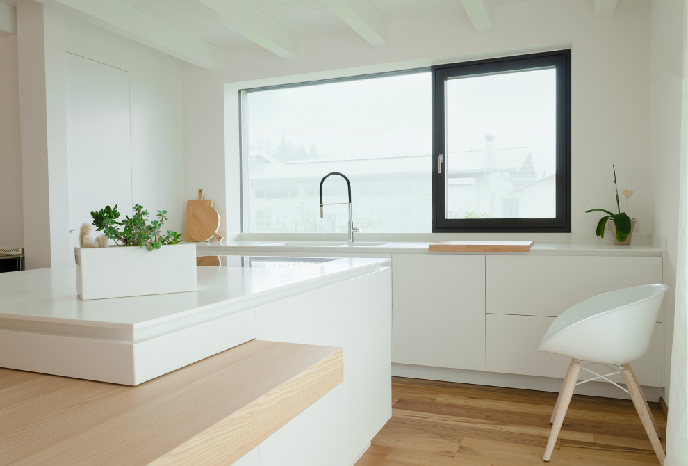 Medium sized scandi galley open plan kitchen in Other with an integrated sink, flat-panel cabinets, white cabinets, composite countertops, black appliances, light hardwood flooring, an island, white worktops and exposed beams.
