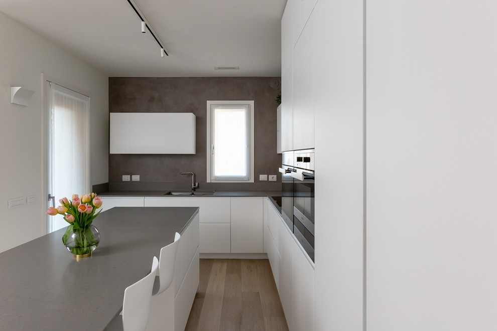 Design ideas for a medium sized modern open plan kitchen in Milan with a double-bowl sink, flat-panel cabinets, white cabinets, engineered stone countertops, black appliances, light hardwood flooring, an island and grey worktops.