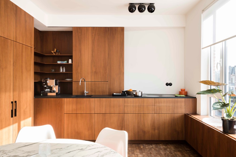 Moderne Wohnküche ohne Insel in L-Form mit hellbraunen Holzschränken, schwarzer Arbeitsplatte, Unterbauwaschbecken, flächenbündigen Schrankfronten, Küchenrückwand in Weiß, Elektrogeräten mit Frontblende und braunem Boden in Mailand