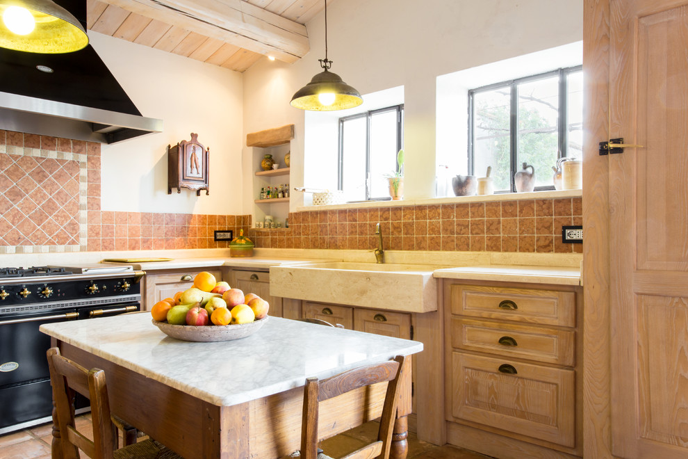 Cette image montre une cuisine américaine rustique en L et bois clair avec un placard avec porte à panneau surélevé, îlot, un évier de ferme, une crédence rouge, un électroménager noir et un plan de travail beige.