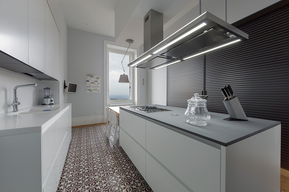Modern inredning av ett grå grått kök, med en integrerad diskho, släta luckor, vita skåp, vitt stänkskydd, glaspanel som stänkskydd, rostfria vitvaror, klinkergolv i keramik, en köksö och flerfärgat golv