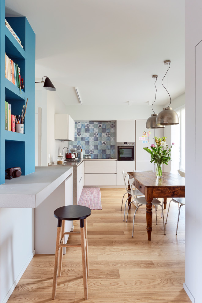 Design ideas for an eclectic l-shaped kitchen/diner in Other with flat-panel cabinets, white cabinets, multi-coloured splashback, stainless steel appliances, light hardwood flooring and beige floors.