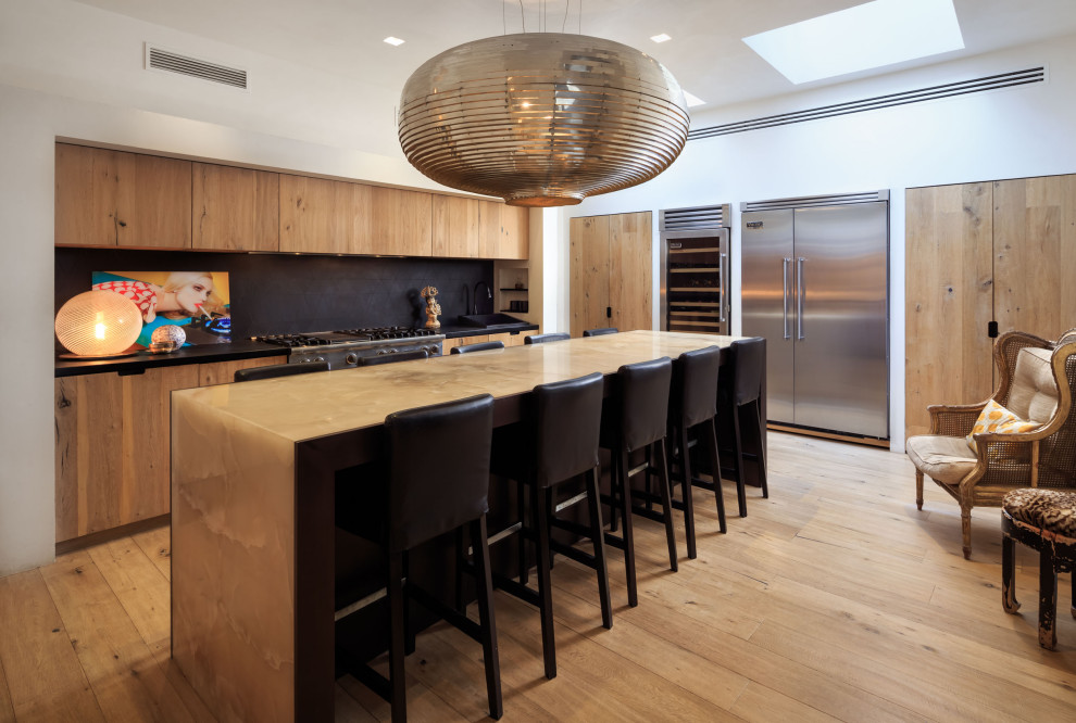 Cette image montre une cuisine design en L et bois brun avec un placard à porte plane, une crédence noire, un électroménager en acier inoxydable, parquet clair, îlot, un sol beige et plan de travail noir.