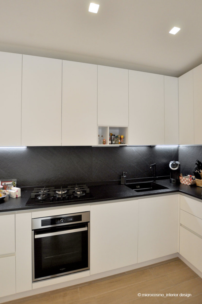Bild på ett litet nordiskt kök, med en nedsänkt diskho, släta luckor, vita skåp, bänkskiva i kvarts, svart stänkskydd, stänkskydd i porslinskakel, svarta vitvaror och klinkergolv i porslin