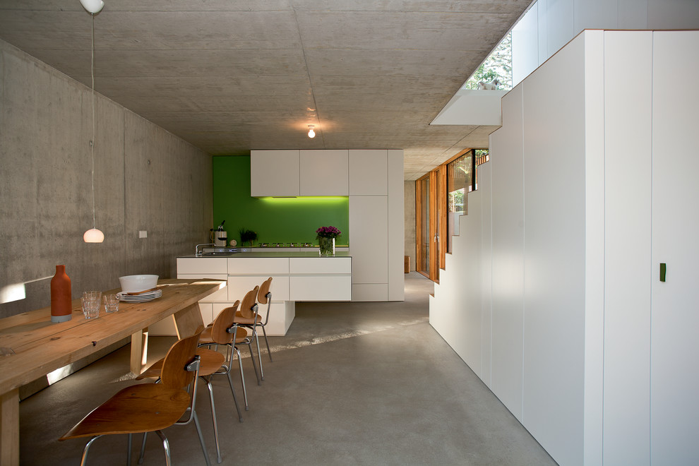 Mittelgroße Moderne Wohnküche mit flächenbündigen Schrankfronten, weißen Schränken, Küchenrückwand in Grün, Betonboden, Kücheninsel, Waschbecken und Elektrogeräten mit Frontblende in Stuttgart