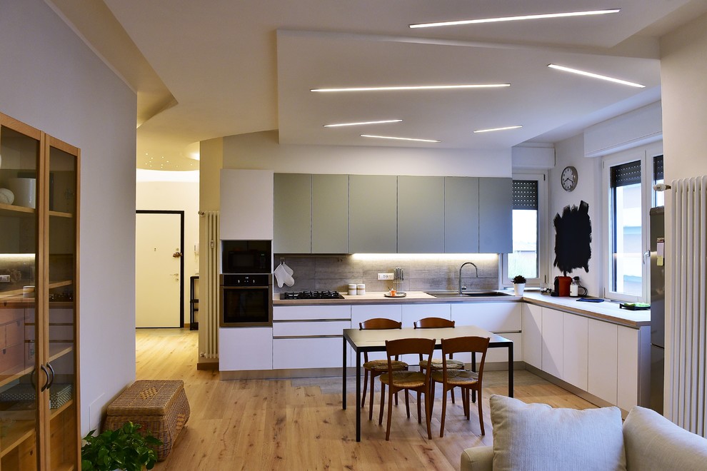 Modern inredning av ett kök, med släta luckor, vita skåp, en dubbel diskho, grått stänkskydd, svarta vitvaror, mellanmörkt trägolv och brunt golv