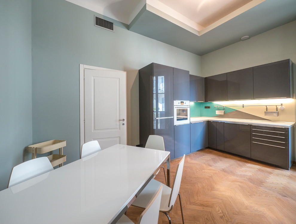 Стильный дизайн: угловая кухня-гостиная среднего размера в современном стиле с накладной мойкой, плоскими фасадами, серыми фасадами, столешницей из ламината, разноцветным фартуком, белой техникой и светлым паркетным полом без острова - последний тренд