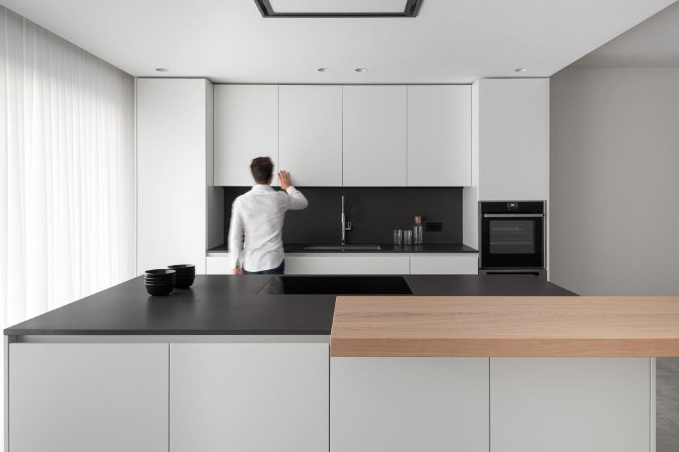 Cette image montre une cuisine parallèle design de taille moyenne avec un évier encastré, un placard à porte plane, des portes de placard blanches, une crédence noire, îlot, un sol gris et plan de travail noir.