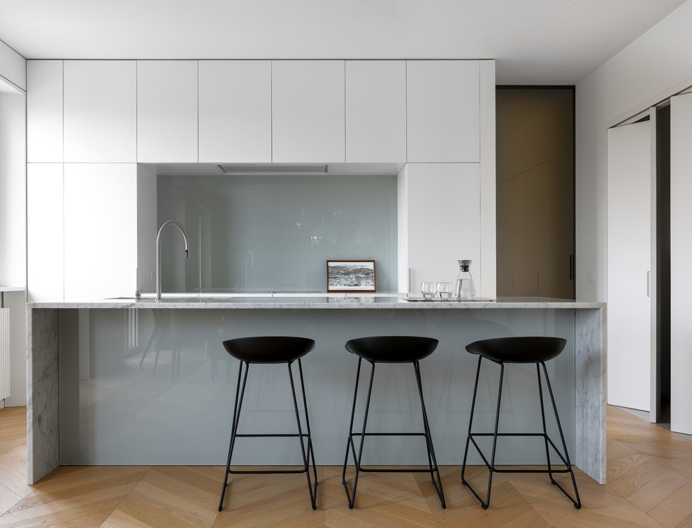 Modern inredning av ett stort vit vitt kök, med en undermonterad diskho, släta luckor, vita skåp, marmorbänkskiva, grått stänkskydd, glaspanel som stänkskydd, mellanmörkt trägolv, en köksö, beiget golv och integrerade vitvaror