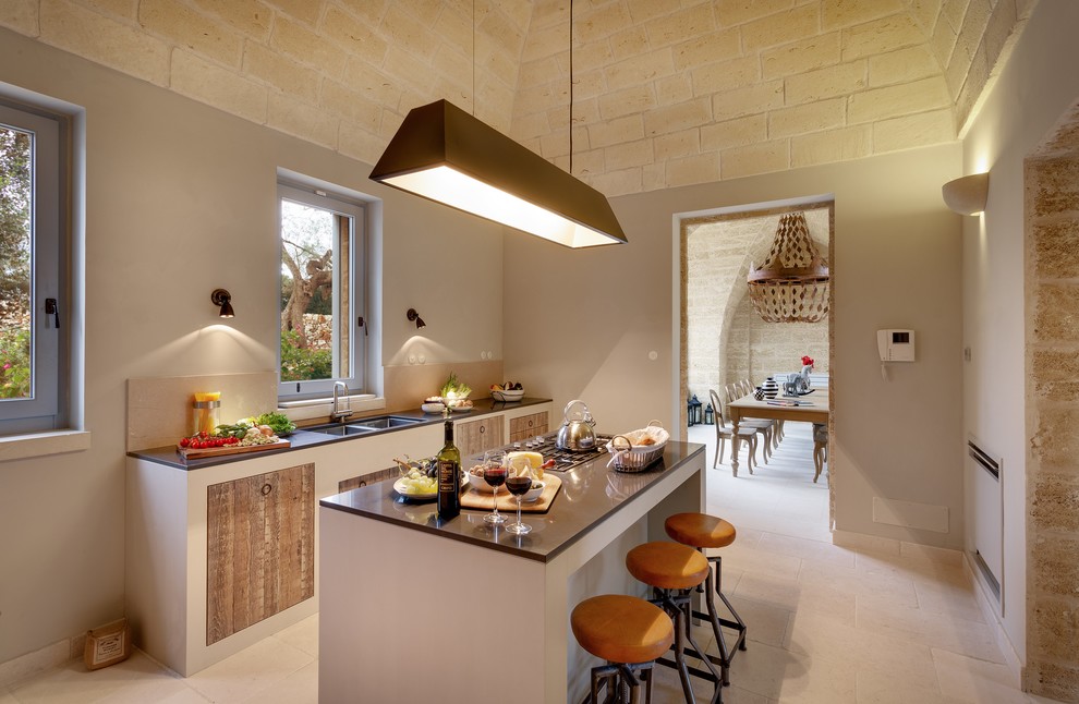 На фото: параллельная кухня в средиземноморском стиле с двойной мойкой, плоскими фасадами, фасадами цвета дерева среднего тона, островом, бежевым полом и серой столешницей