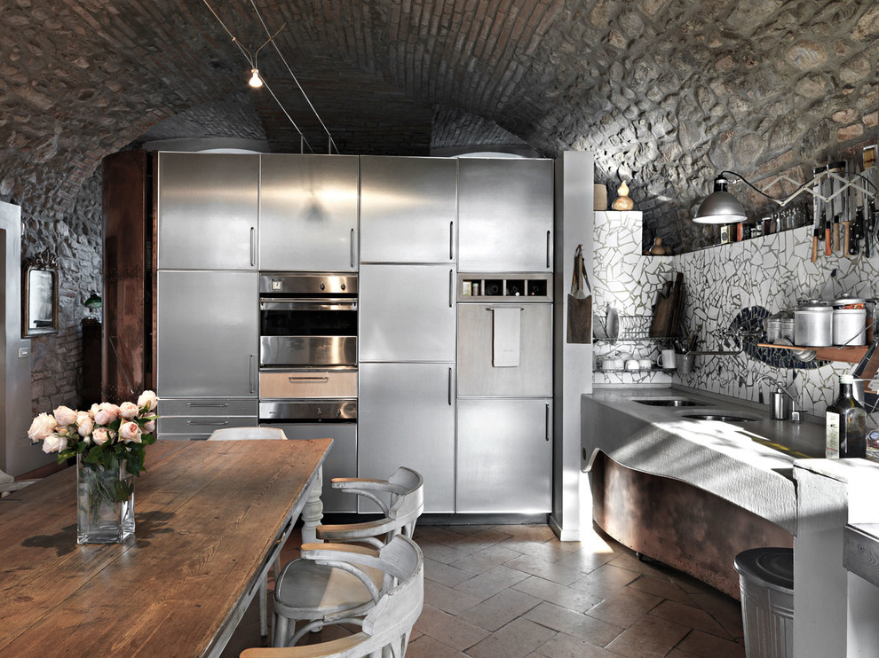 Inspiration for an industrial l-shaped kitchen in Other with a double-bowl sink, stainless steel cabinets, flat-panel cabinets, mosaic tiled splashback, stainless steel appliances, brick flooring, no island, brown floors and grey worktops.