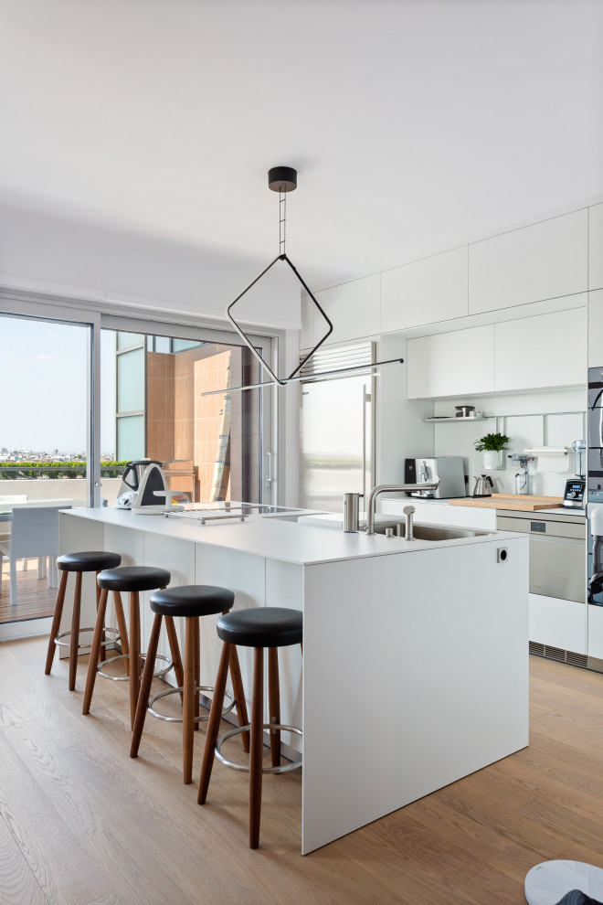 Inspiration for a large modern galley open plan kitchen in Milan with a built-in sink, flat-panel cabinets, white splashback, glass sheet splashback, stainless steel appliances, light hardwood flooring, an island and white worktops.