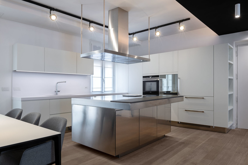 Large contemporary l-shaped kitchen/diner in Rome with a submerged sink, flat-panel cabinets, white cabinets, white splashback, light hardwood flooring, an island, stainless steel worktops, integrated appliances, brown floors and grey worktops.