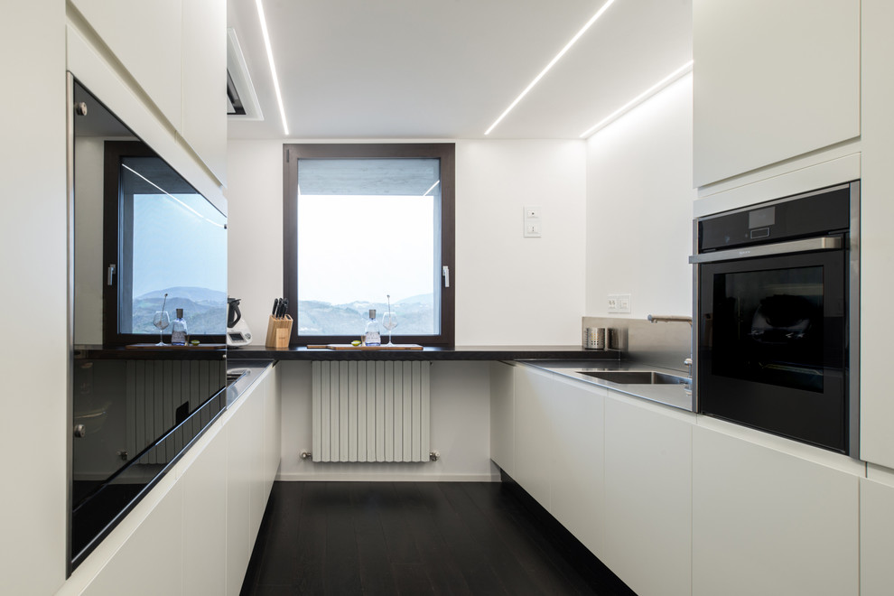 Design ideas for a medium sized modern u-shaped kitchen/diner in Other with an integrated sink, flat-panel cabinets, white cabinets, stainless steel worktops, grey splashback, black appliances, dark hardwood flooring, no island and black floors.