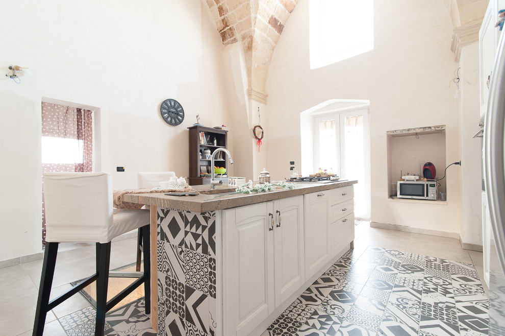 Immagine di un cucina con isola centrale mediterraneo con lavello sottopiano, ante con bugna sagomata, ante bianche, top in marmo, pavimento in cementine e pavimento beige