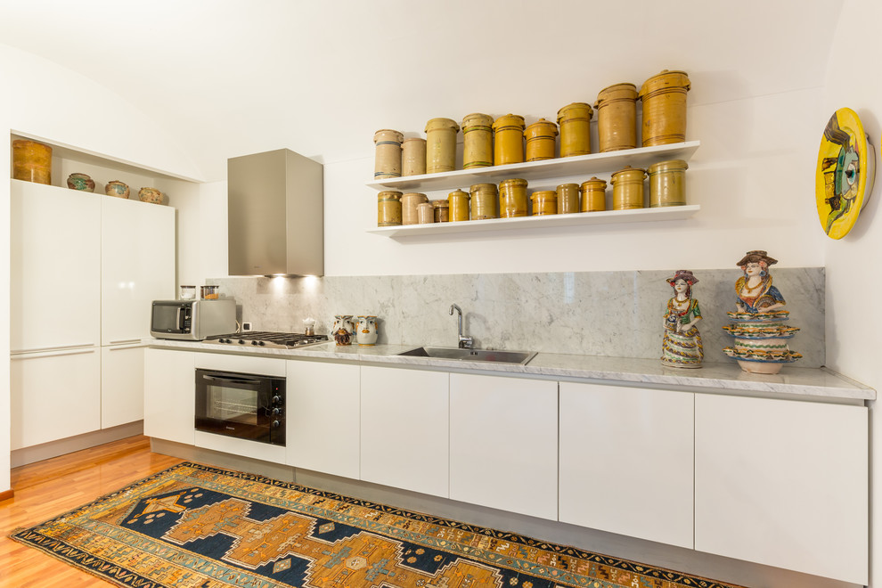 Mediterranean single-wall kitchen in Catania-Palermo with a built-in sink, flat-panel cabinets, white cabinets, marble worktops, grey splashback, stone slab splashback, black appliances and light hardwood flooring.