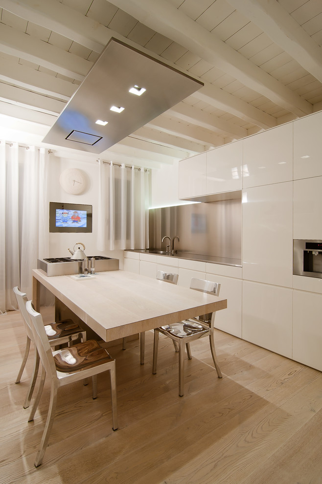 Modern inredning av ett kök, med släta luckor, vita skåp, bänkskiva i rostfritt stål, stänkskydd med metallisk yta, rostfria vitvaror, ljust trägolv, en köksö och beiget golv