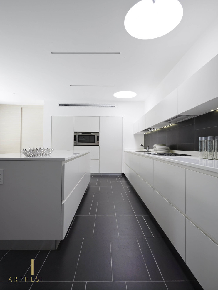 Cette image montre une cuisine minimaliste avec un évier 1 bac, des portes de placard blanches, un plan de travail en quartz modifié, un électroménager en acier inoxydable, un sol en ardoise et îlot.