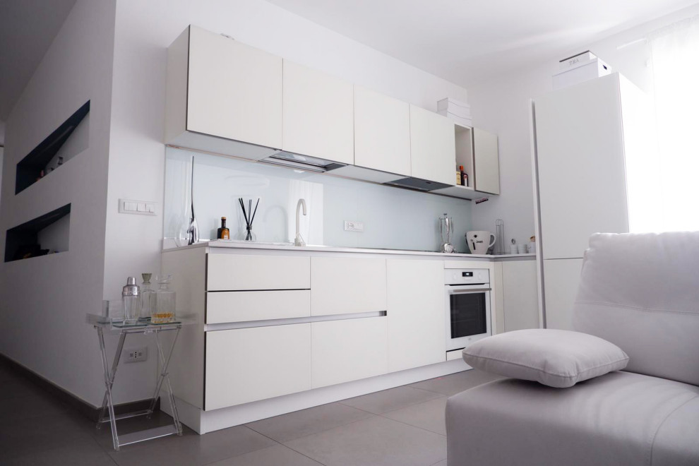 Bild på ett litet vit linjärt vitt kök med öppen planlösning, med en integrerad diskho, vita skåp, bänkskiva i glas, vitt stänkskydd, rostfria vitvaror, klinkergolv i porslin och brunt golv