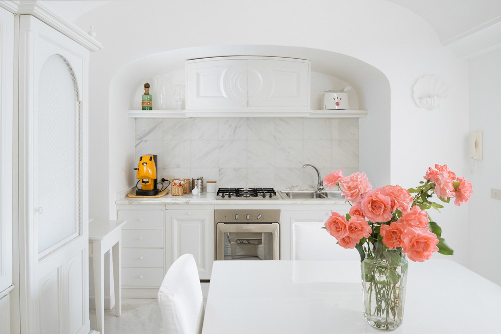 Diseño de cocina lineal mediterránea pequeña abierta con fregadero encastrado, puertas de armario blancas, encimera de mármol, salpicadero blanco, salpicadero de losas de piedra y electrodomésticos de acero inoxidable