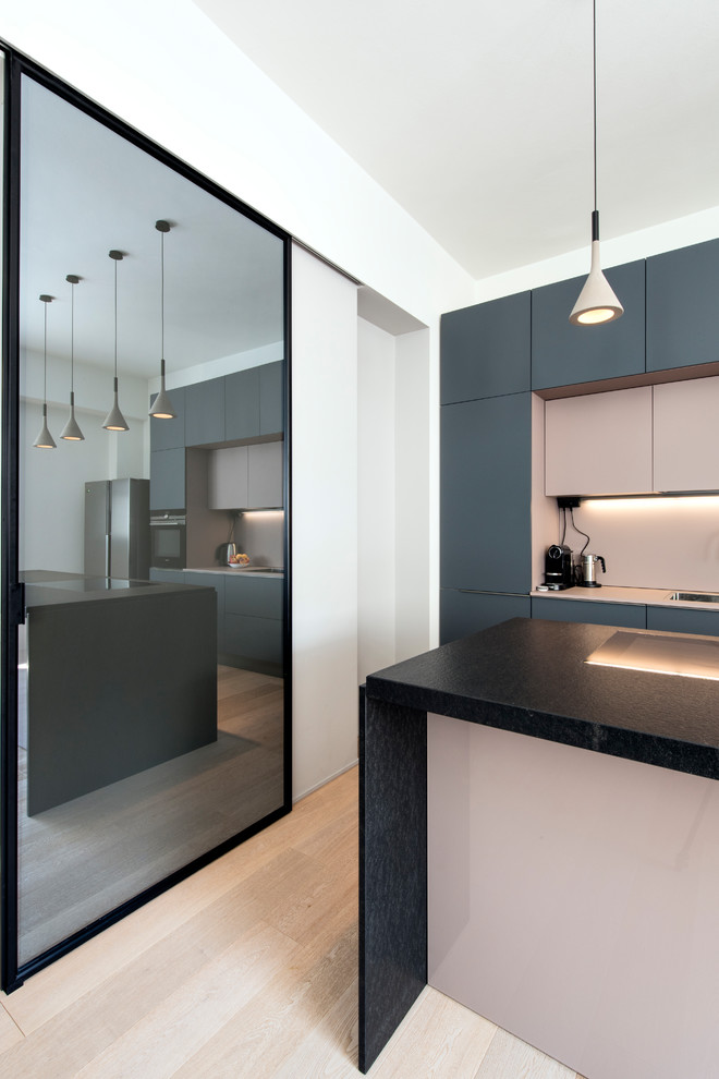 Photo of a large contemporary galley open plan kitchen in Milan with a built-in sink, flat-panel cabinets, grey cabinets, marble worktops, pink splashback, glass sheet splashback, black appliances, light hardwood flooring, an island, beige floors and black worktops.