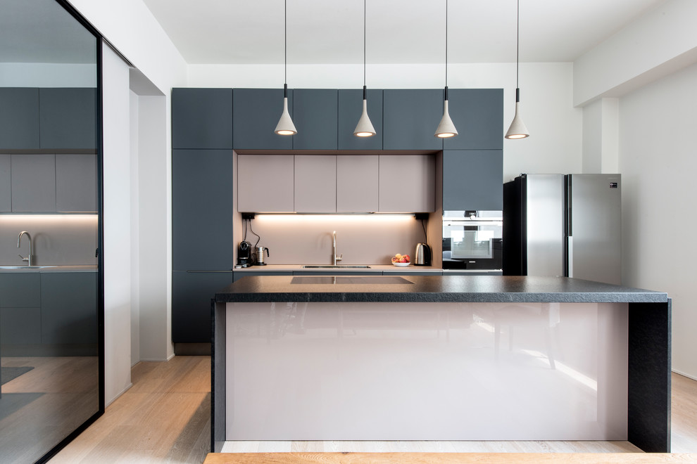 Design ideas for a large contemporary galley open plan kitchen in Milan with flat-panel cabinets, black appliances, light hardwood flooring, an island, black worktops, a built-in sink, grey cabinets, marble worktops, pink splashback, glass sheet splashback and beige floors.