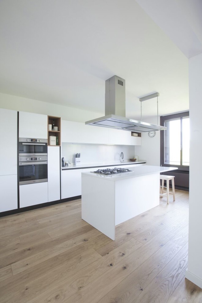 Photo of a large contemporary galley enclosed kitchen in Milan with a built-in sink, flat-panel cabinets, white cabinets, engineered stone countertops, grey splashback, stainless steel appliances, medium hardwood flooring, an island and brown floors.
