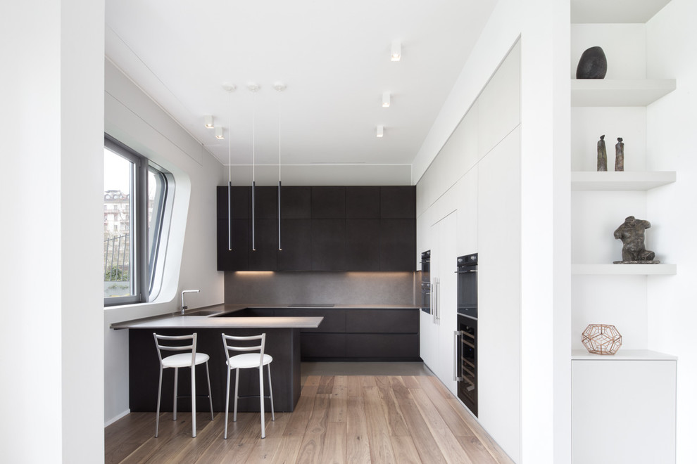 Cette image montre une cuisine minimaliste en U avec un placard à porte plane, des portes de placard noires, une crédence grise, une péninsule, un sol en bois brun, un sol marron et un plan de travail gris.
