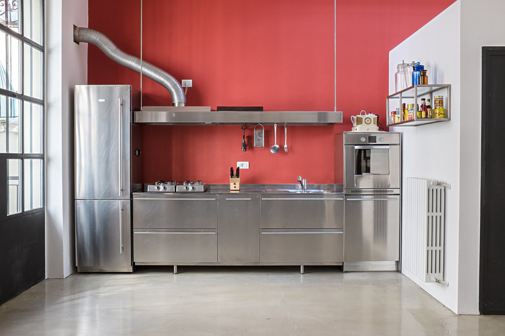 ミラノにある広いインダストリアルスタイルのおしゃれなI型キッチン (フラットパネル扉のキャビネット、ステンレスキャビネット、ステンレスカウンター、赤いキッチンパネル、シルバーの調理設備) の写真
