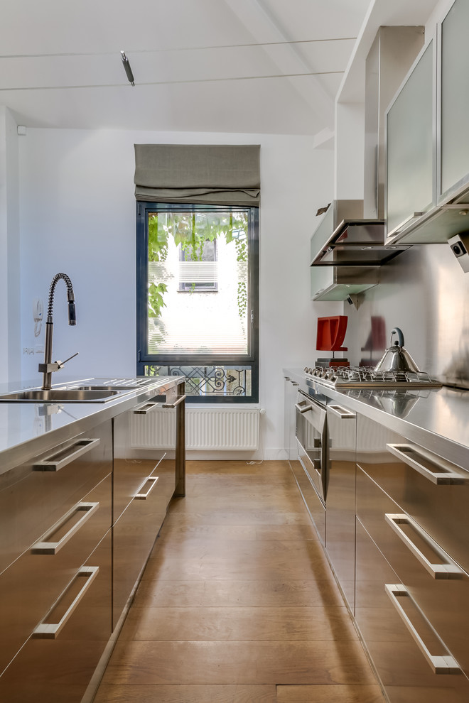 Bild på ett funkis kök, med en integrerad diskho, skåp i rostfritt stål, bänkskiva i rostfritt stål, rostfria vitvaror, ljust trägolv och flera köksöar
