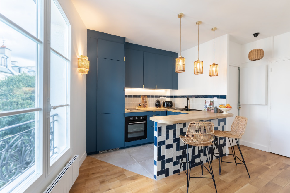 Foto di una cucina ad U design con lavello da incasso, ante lisce, ante blu, top in legno, paraspruzzi bianco, elettrodomestici neri, penisola, pavimento grigio e top beige