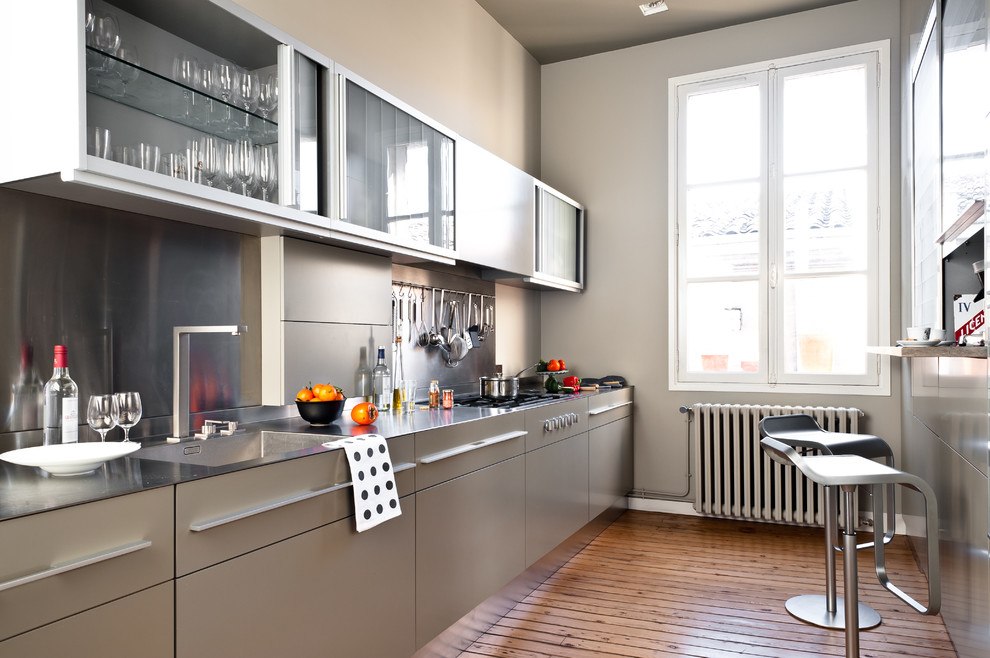 Modelo de cocina contemporánea, de tamaño medio, cerrada, sin isla, con fregadero integrado, armarios con paneles lisos, puertas de armario grises, encimera de acero inoxidable, salpicadero metalizado, suelo de madera en tonos medios y barras de cocina