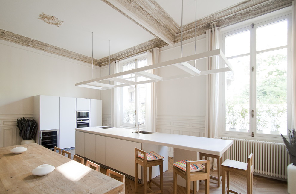 Große Moderne Wohnküche mit Unterbauwaschbecken, flächenbündigen Schrankfronten, weißen Schränken, Küchengeräten aus Edelstahl, braunem Holzboden und Kücheninsel in Paris